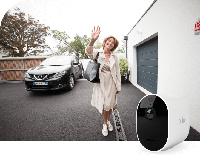 Arlo-valvontakamera ja nainen seisoo talon pihalla hymyillen ja heilauttaen laukku kädessä.