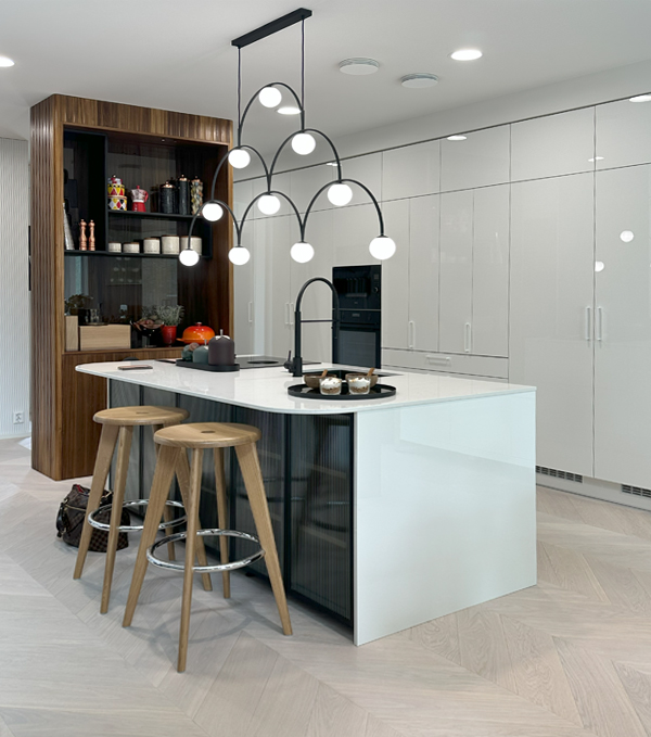 Näkymä Oulun Merta Mainingin keittiöön. Pääosin valkoisen keittiön kaapit on upotettu seinään, pinnat ovat kiiltäviä ja keittiösaarekkeen päällä roikkuu 20-lukua henkivä musta kerroksellinen valaisin, jossa on vaaleita polttimoita. Taustalla on ruskea avokaappi.