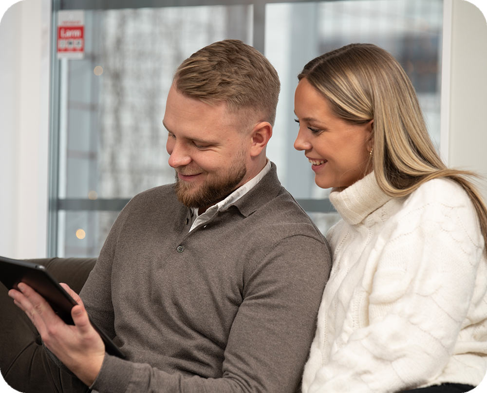 Pariskunta istuu sohvalla ja katsoo hymyillen tablettia, taustalla ikkuna ja Verisuren tarra.